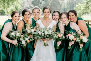 Bride dressed in a white dress and surrounded by bridesmaids in emerald green. All are holding floral bouquets at All4One Farms wedding venue.