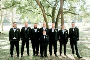 The groom is surrounded by his groomsmen. All are dressed in black tuxedos on the front lawn of All4One Farms.