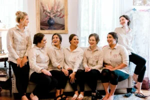 Bridesmaids dressed in matching casual shirts gather around the bride in the bridal suite at All4One Farms wedding venue.