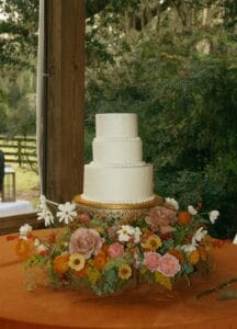 A white three-tiered wedding cake with fresh florals around the base in the reception barn at All4One Farms. The flowers are oranges and pinks and yellows. 