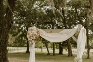 Wooden arch decorated with drapping and white and peach florals under the shade of giant oak trees.